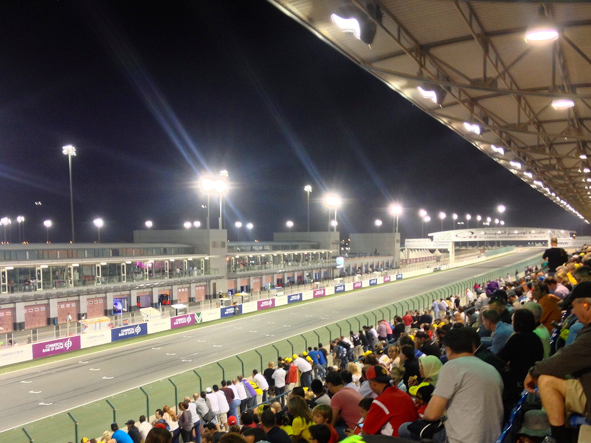 Crowd in the covered main grandstand at Lusail International Circuit at night, looking down the pit straight